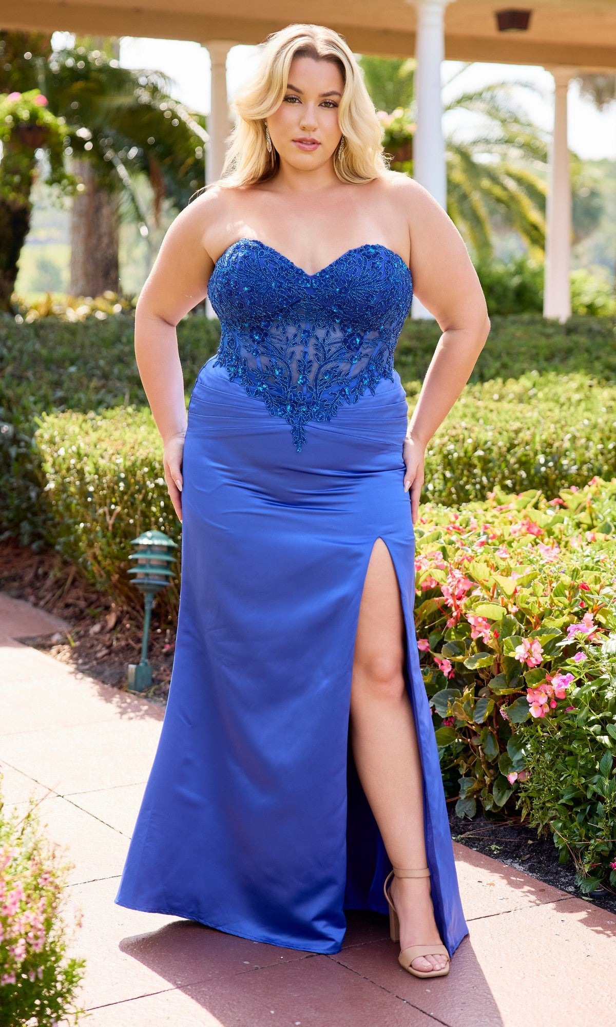 A woman stands outdoors on a garden path wearing the Long Plus-Size Prom Dress Primavera 14109 in royal blue, featuring a strapless design with a sheer embroidered bodice and satin sheath skirt.